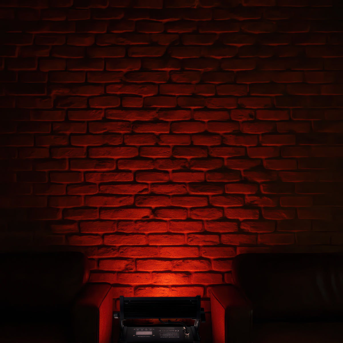Ein roter Bühnenscheinwerfer beleuchtet eine gemauerte Wand von unten, flankiert von Sesseln.