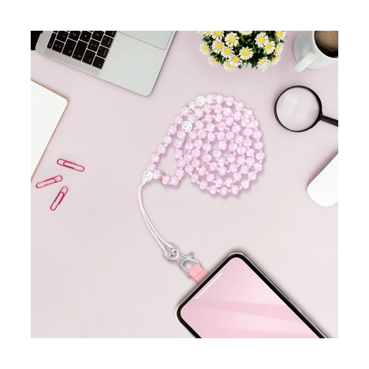 Ein rosafarbenes Perlen-Handyband an einem Smartphone auf einem Schreibtisch mit Laptop und Blumen.