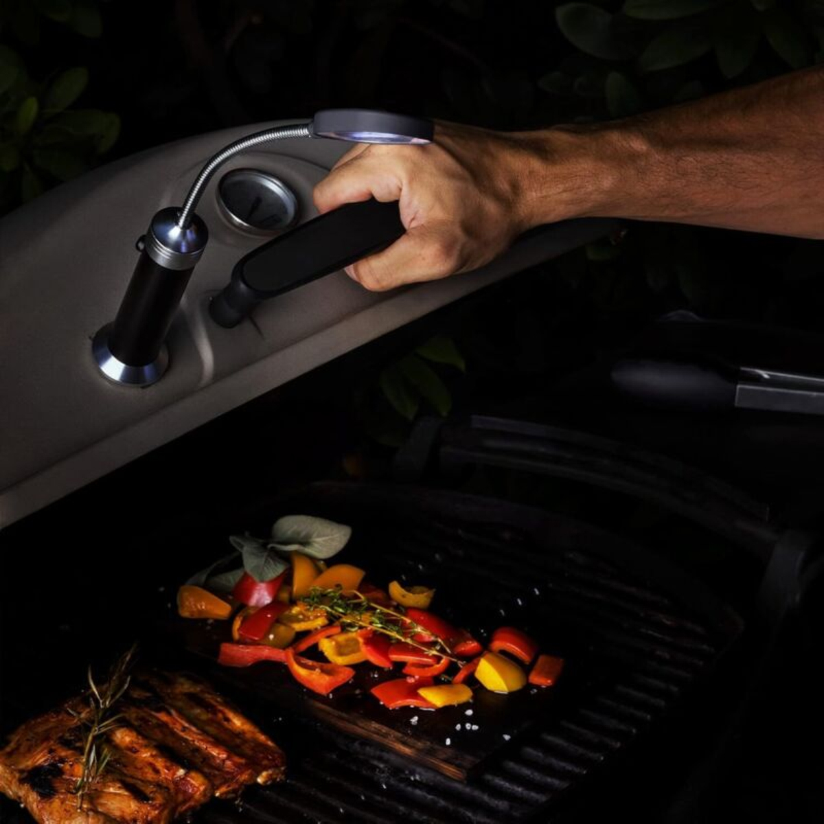 Eine Hand öffnet den Grilldeckel, beleuchtet von einer flexiblen Grilllampe; Gemüseschnitze und Fleisch braten auf dem Rost.