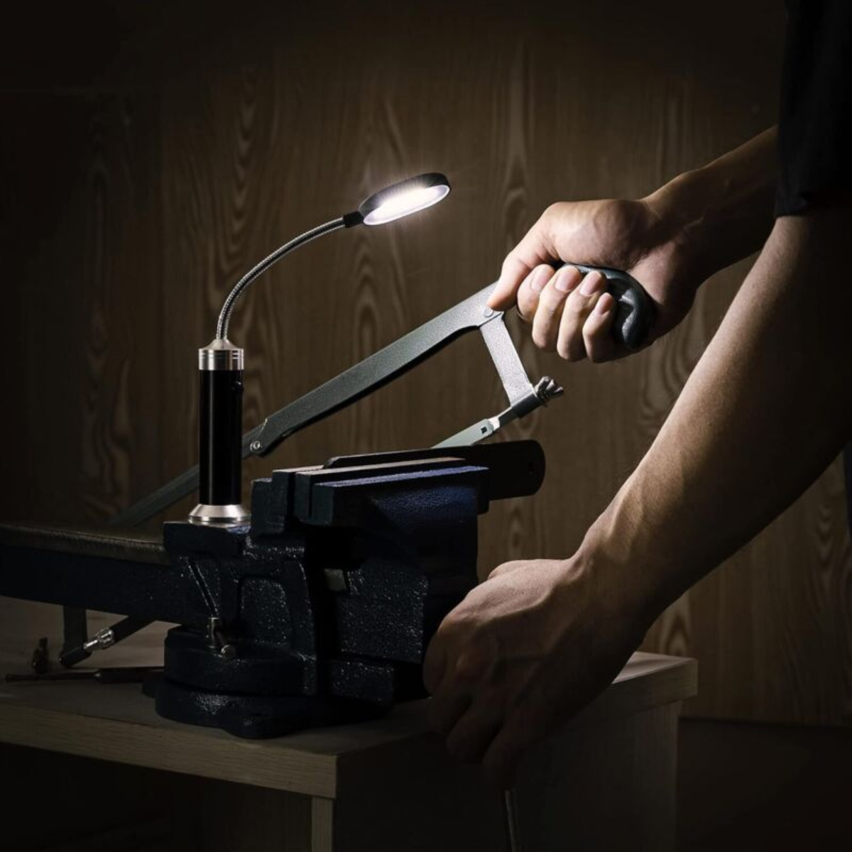 Eine Hand sägt Holz in einem Schraubstock, beleuchtet von einer kleinen Lampe auf einem hölzernen Tisch.
