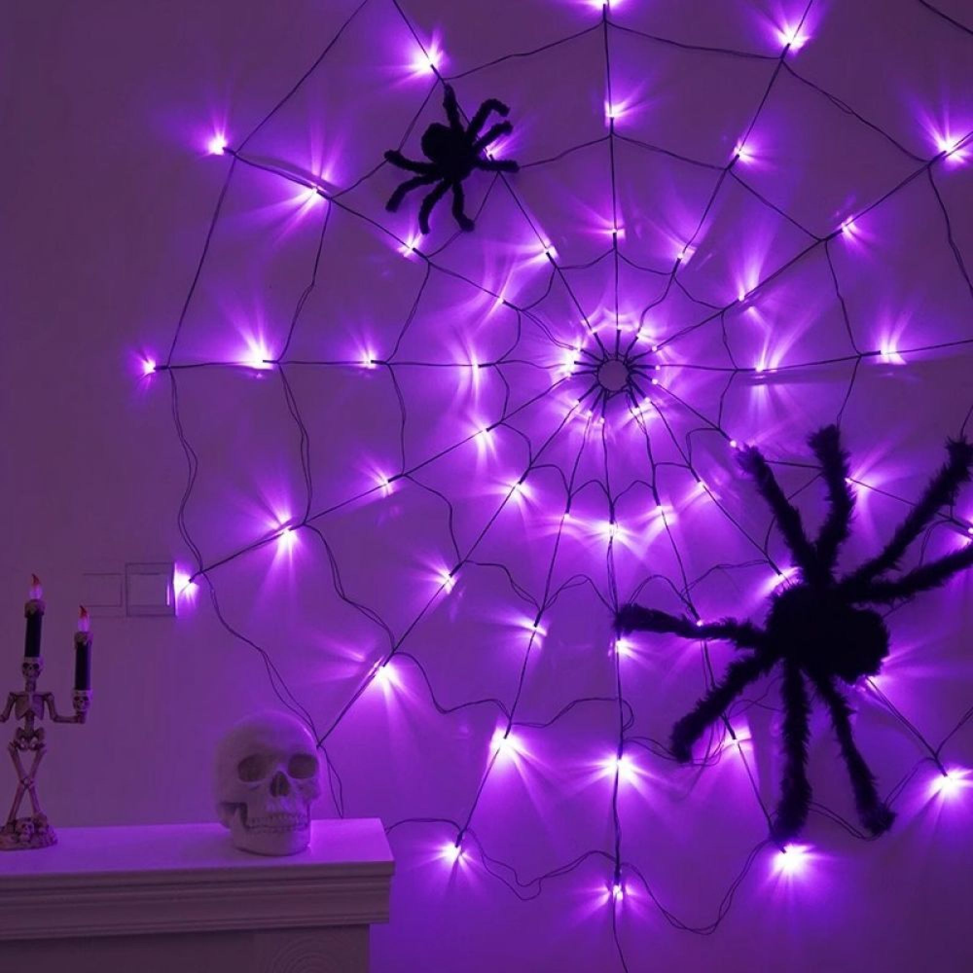 Eine leuchtend lila LED-Spinnennetz-Dekoration mit zwei schwarzen Spinnen an einer Wand, neben einem Totenkopf und Kerzen.