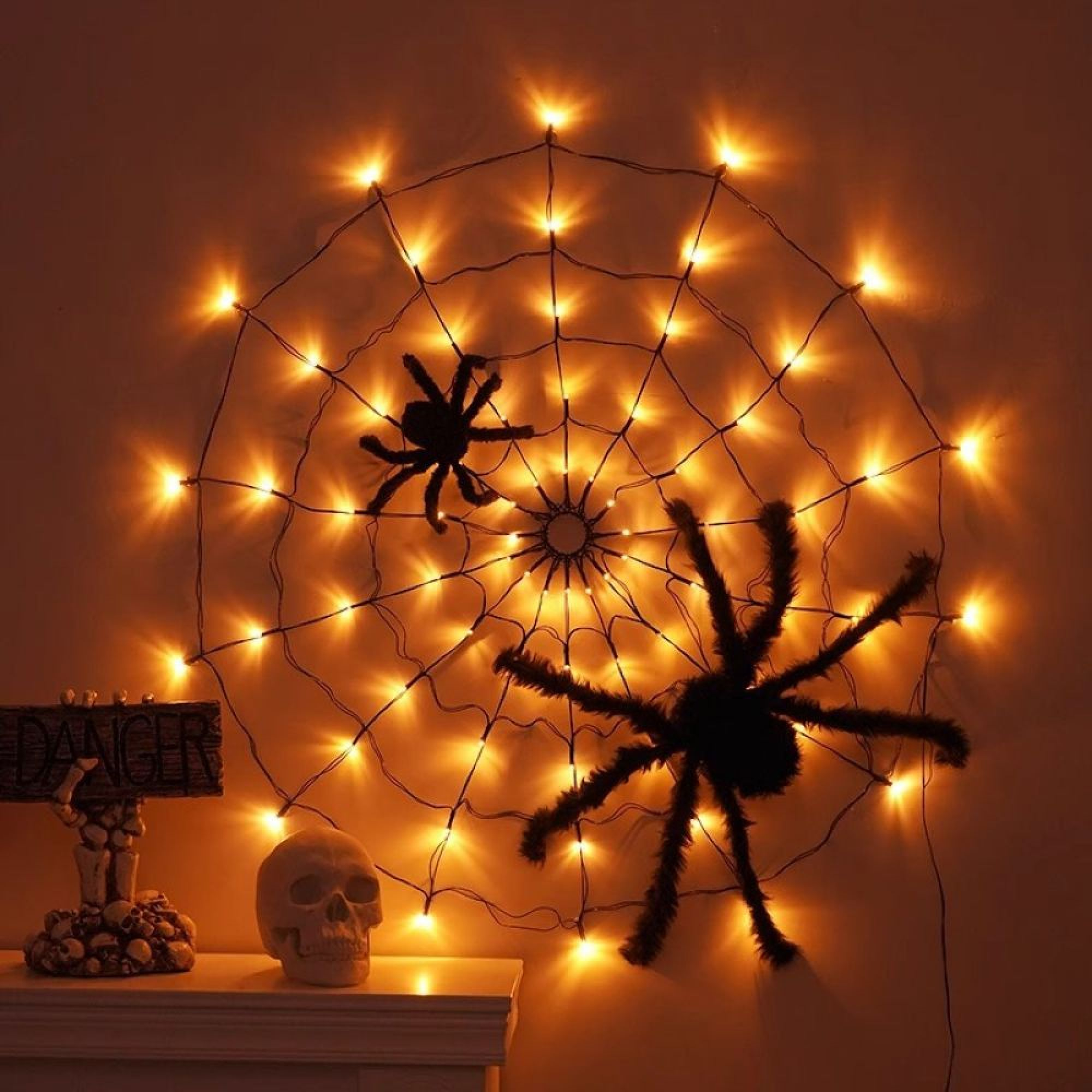 Eine leuchtende Spinnennetz-Lichterkette mit zwei schwarzen Spinnen an einer Wand, daneben ein Totenkopf.