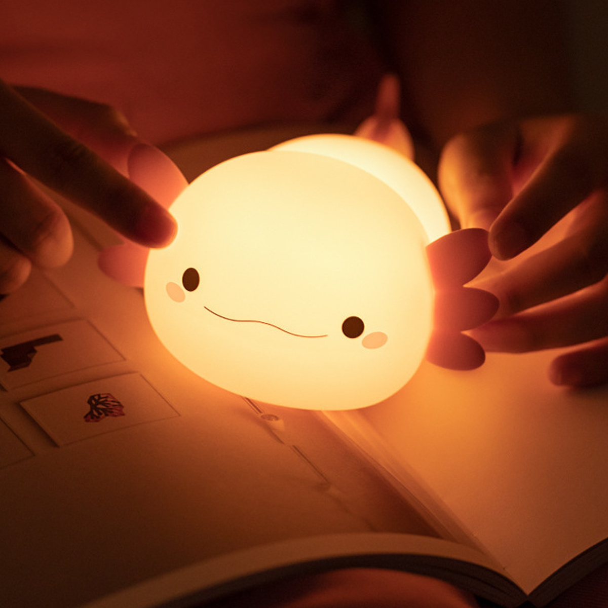 Ein süßes, leuchtendes Axolotl-Nachtlicht ruht auf einem offenen Buch, gehalten von Händen.