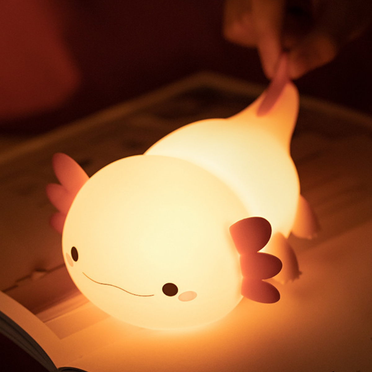 Ein leuchtendes rosa Axolotl-Silikon-Nachtlicht auf einem offenen Buch, während eine Hand seinen Schwanz hält.