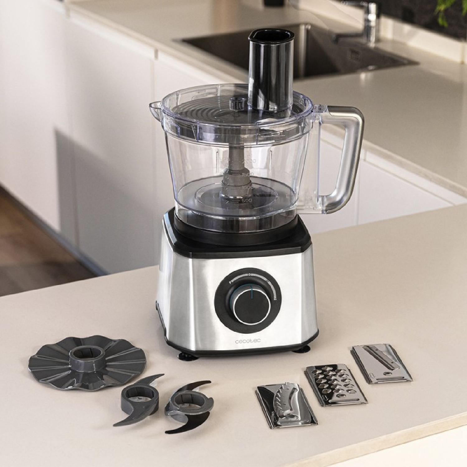 Un robot da cucina argentato con ciotola trasparente e accessori su un bancone bianco.