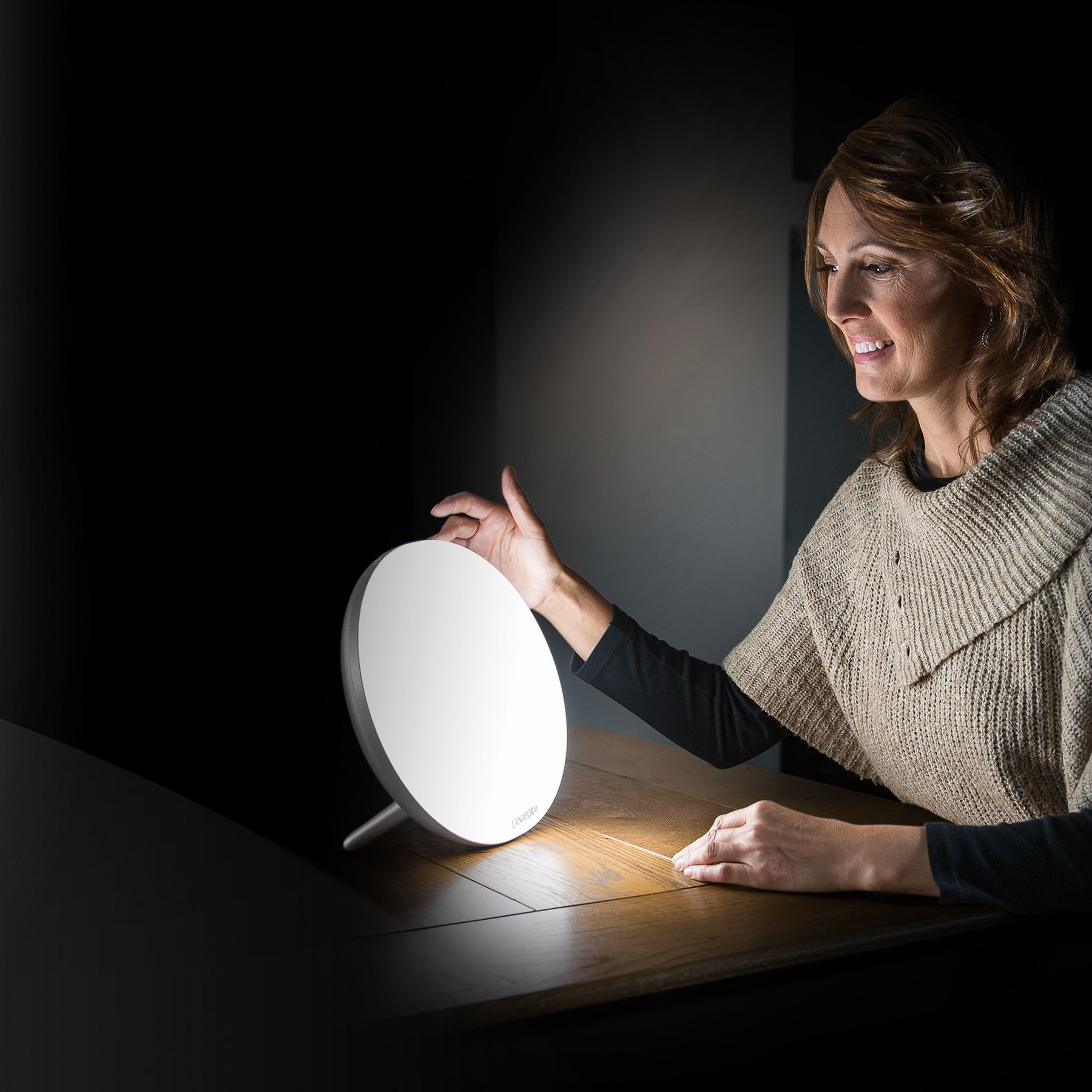 Een vrouw zit aan een houten tafel en bedient een heldere, ronde daglichtlamp in een donkere kamer.