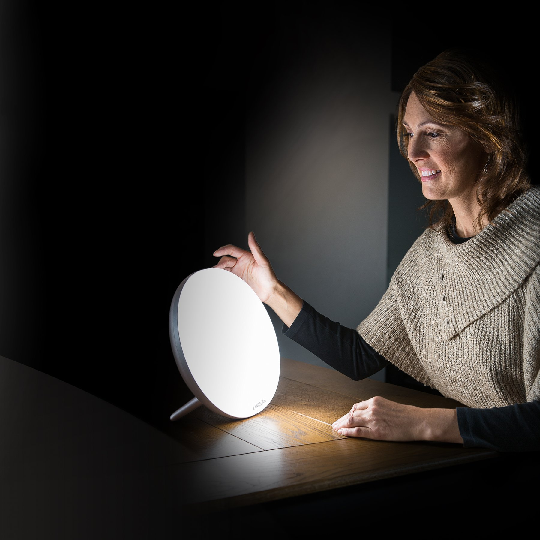 Een vrouw zit aan een houten tafel en kijkt naar een heldere, ronde daglichtlamp voor haar.