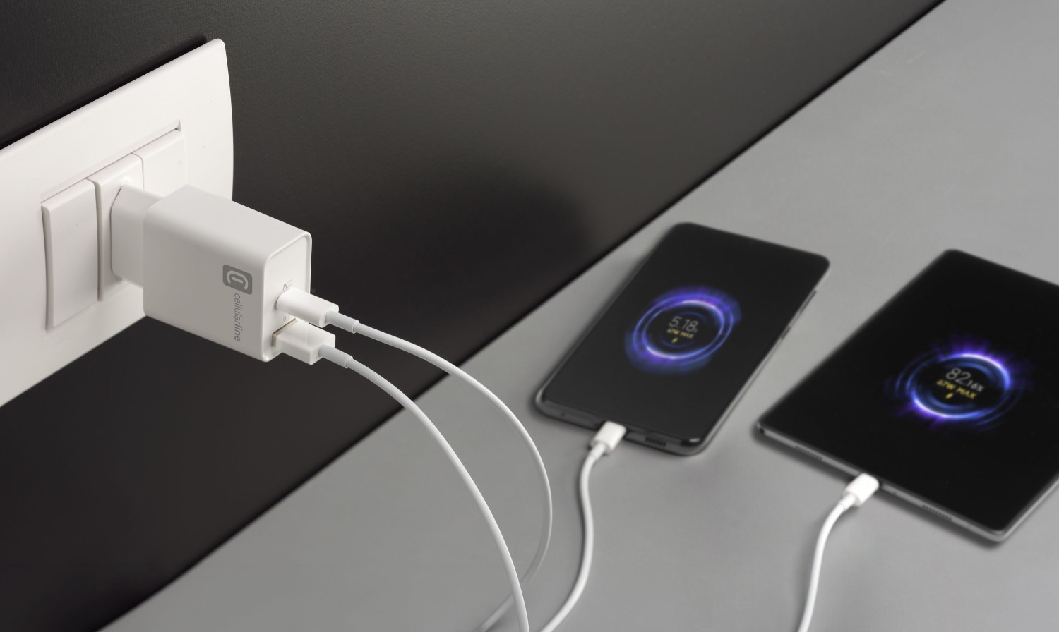 Un cargador de pared USB blanco enchufado carga un teléfono y una tableta en una superficie gris.