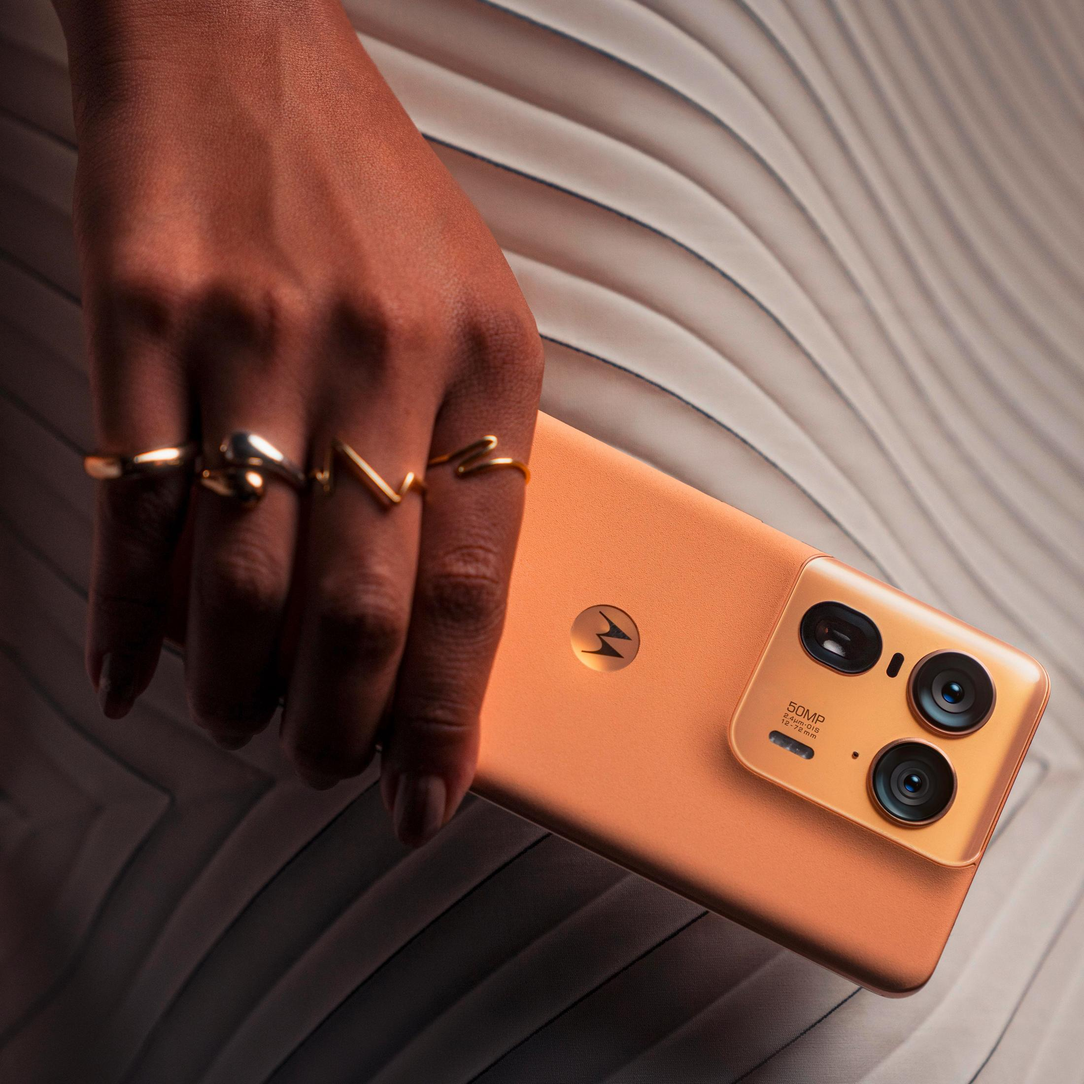 Una mano con anelli tiene uno smartphone Motorola arancione su uno sfondo strutturato.
