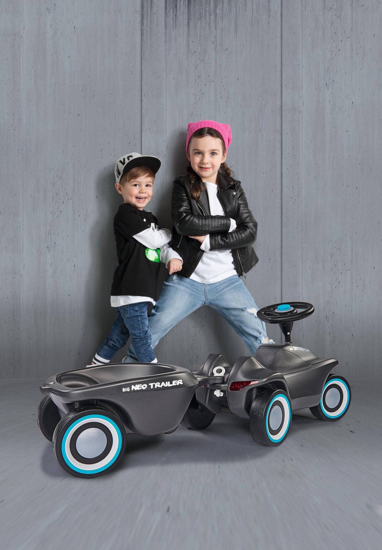 Zwei glückliche Kinder stehen hinter einem schwarzen Rutschauto mit Anhänger vor grauer Wand.