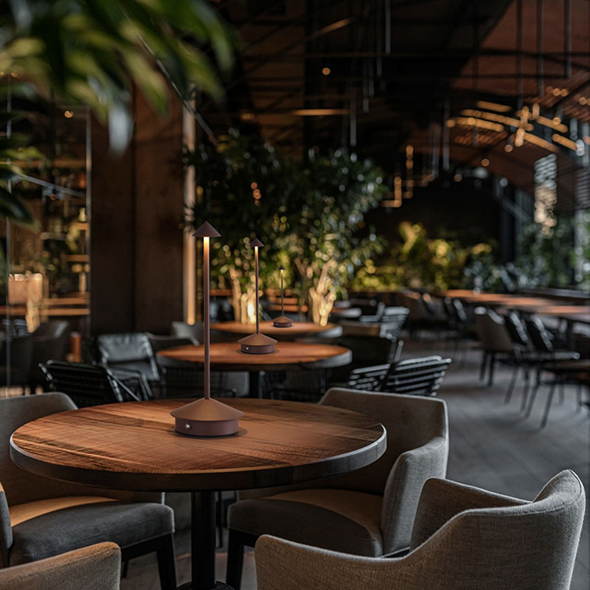 Moderne, schlanke Lampen stehen auf runden Holztischen in einem dunklen Restaurant mit gepolsterten Stühlen und Pflanzen.