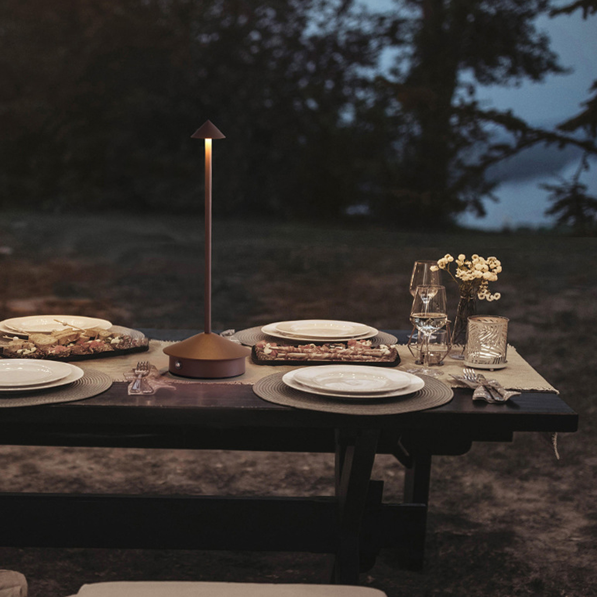 Ein beleuchteter Esstisch im Freien bei Dämmerung mit Tellern, Gläsern, Blumen und einer schlanken Lampe.