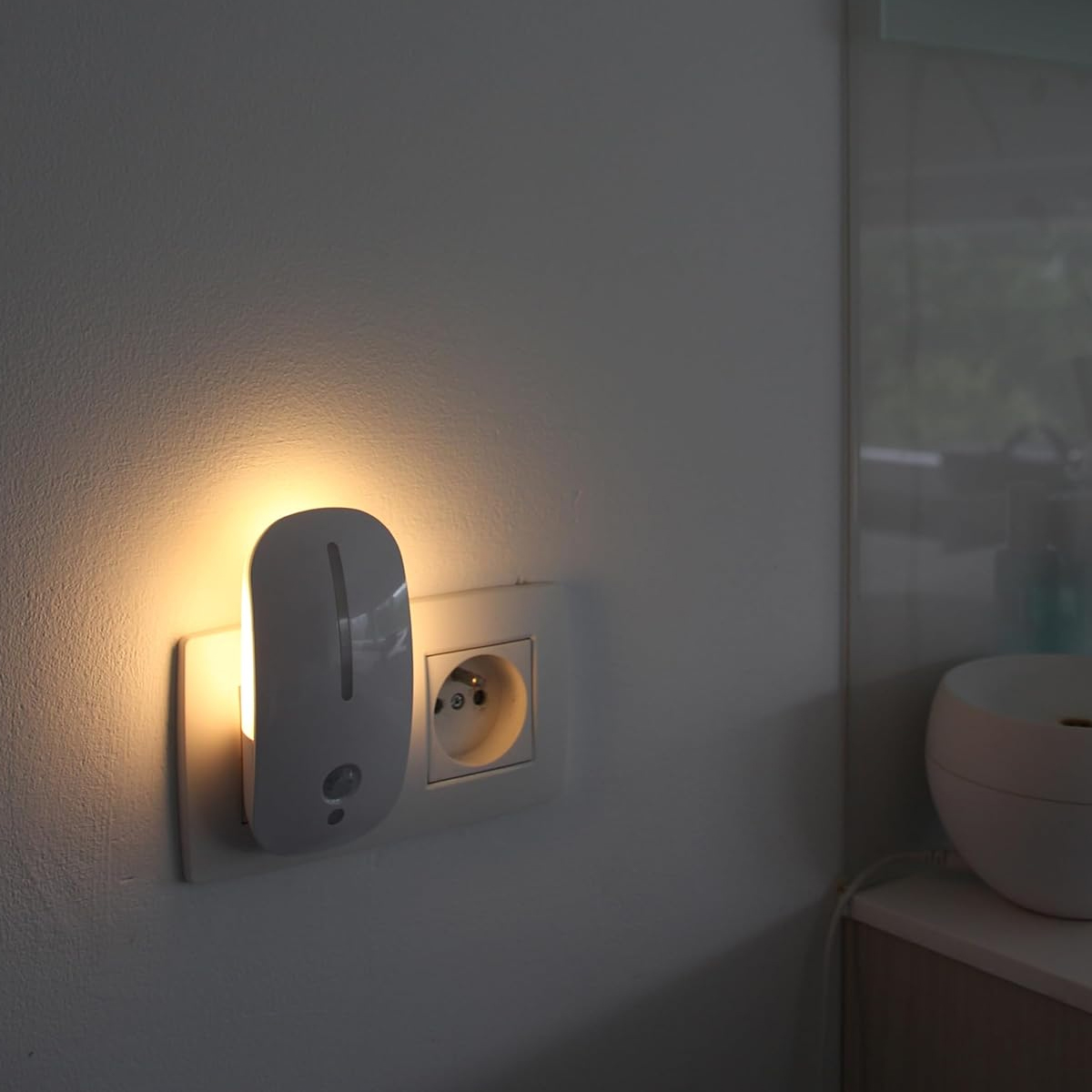 Eine weiße Steckdosenleuchte strahlt warmes Licht aus, neben einer Steckdose an einer Wand.