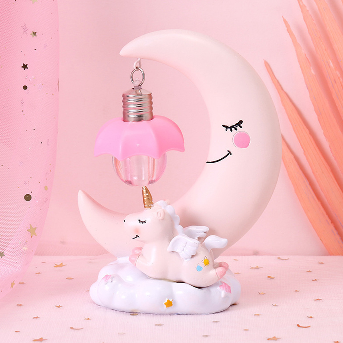 Eine rosafarbene Mondlampe mit einer hängenden Glühbirne und einem Einhorn auf einer weißen Wolke vor rosafarbenem Hintergrund.
