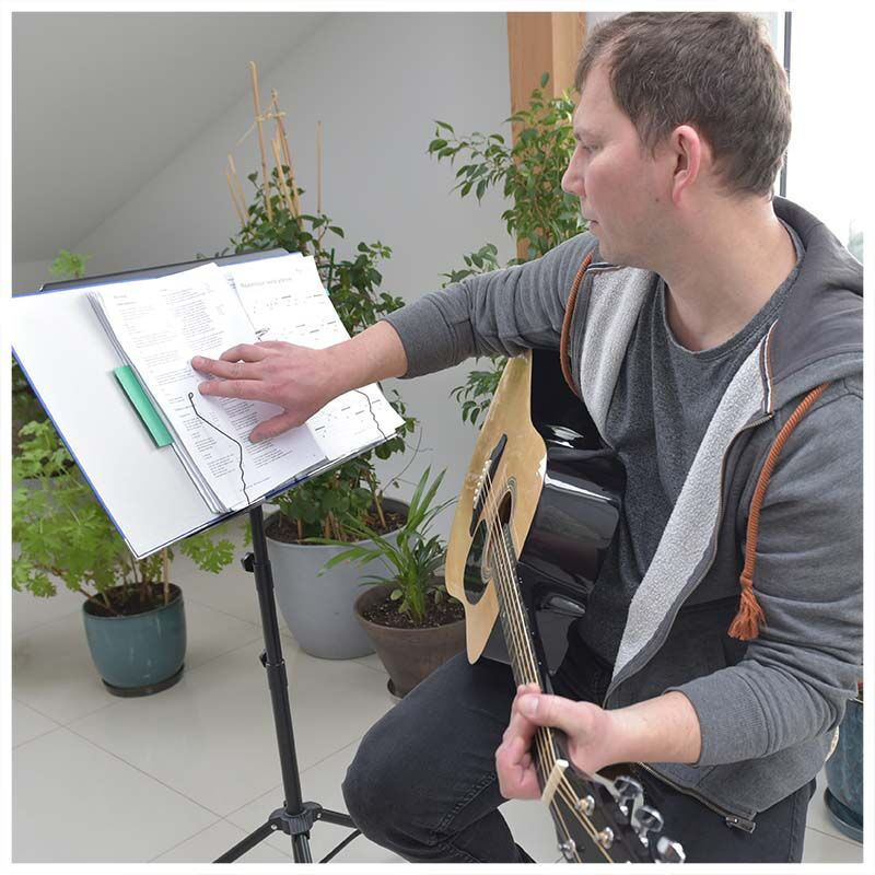 Ein Mann im grauen Hoodie spielt Akustikgitarre und zeigt auf Noten auf einem Ständer, im Hintergrund Topfpflanzen.