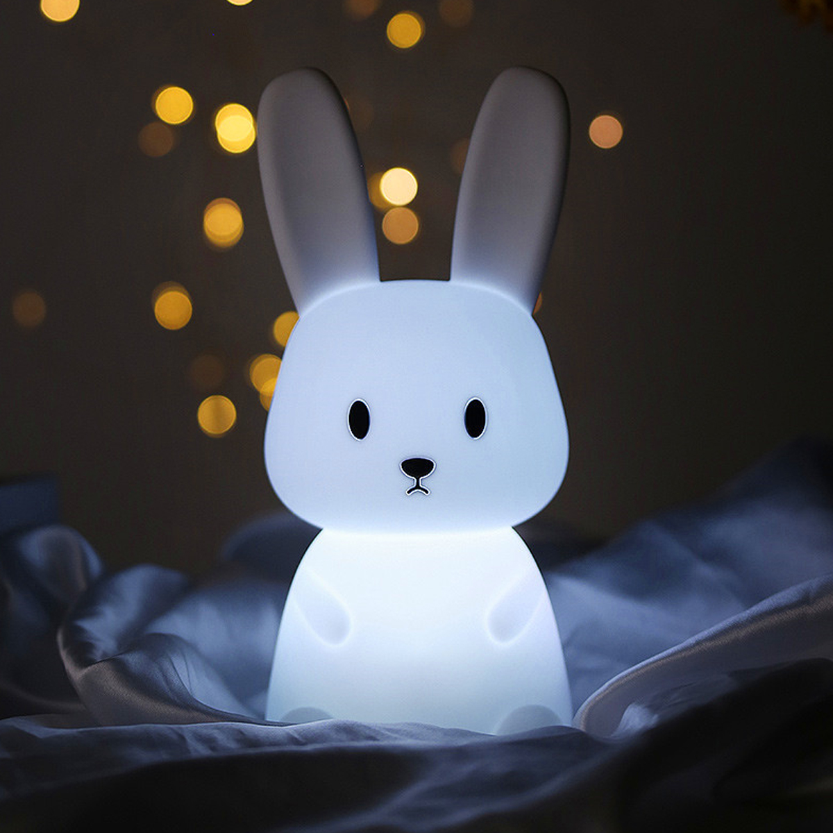 Eine süße, weiß leuchtende Hasenlampe steht auf zerknitterten, bläulichen Laken vor einem warmen, verschwommenen Hintergrund.