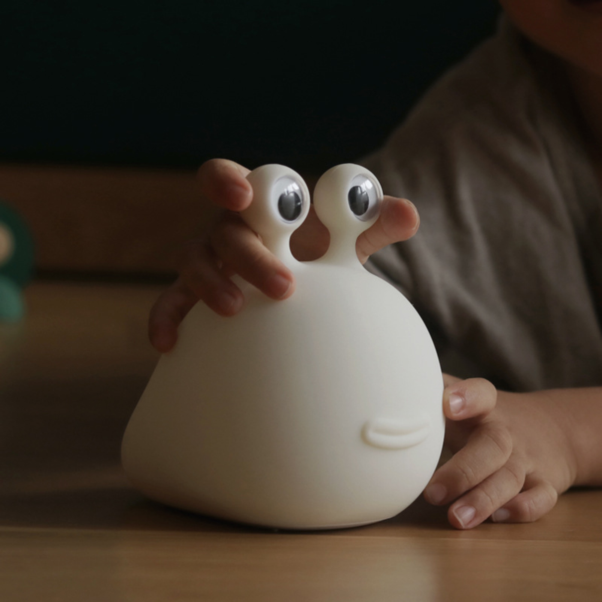 Kinderhände halten sanft eine weiße, schneckenförmige Silikonlampe mit Wackelaugen auf einem Holztisch.