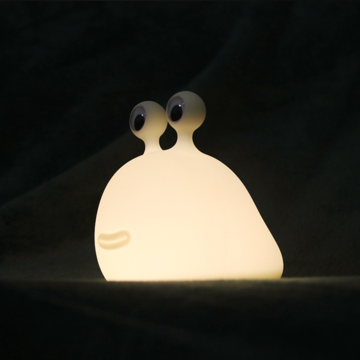 Eine leuchtende weiße Schneckenlampe aus Silikon mit Wackelaugen steht auf dunklem Grund vor dunklem Hintergrund.