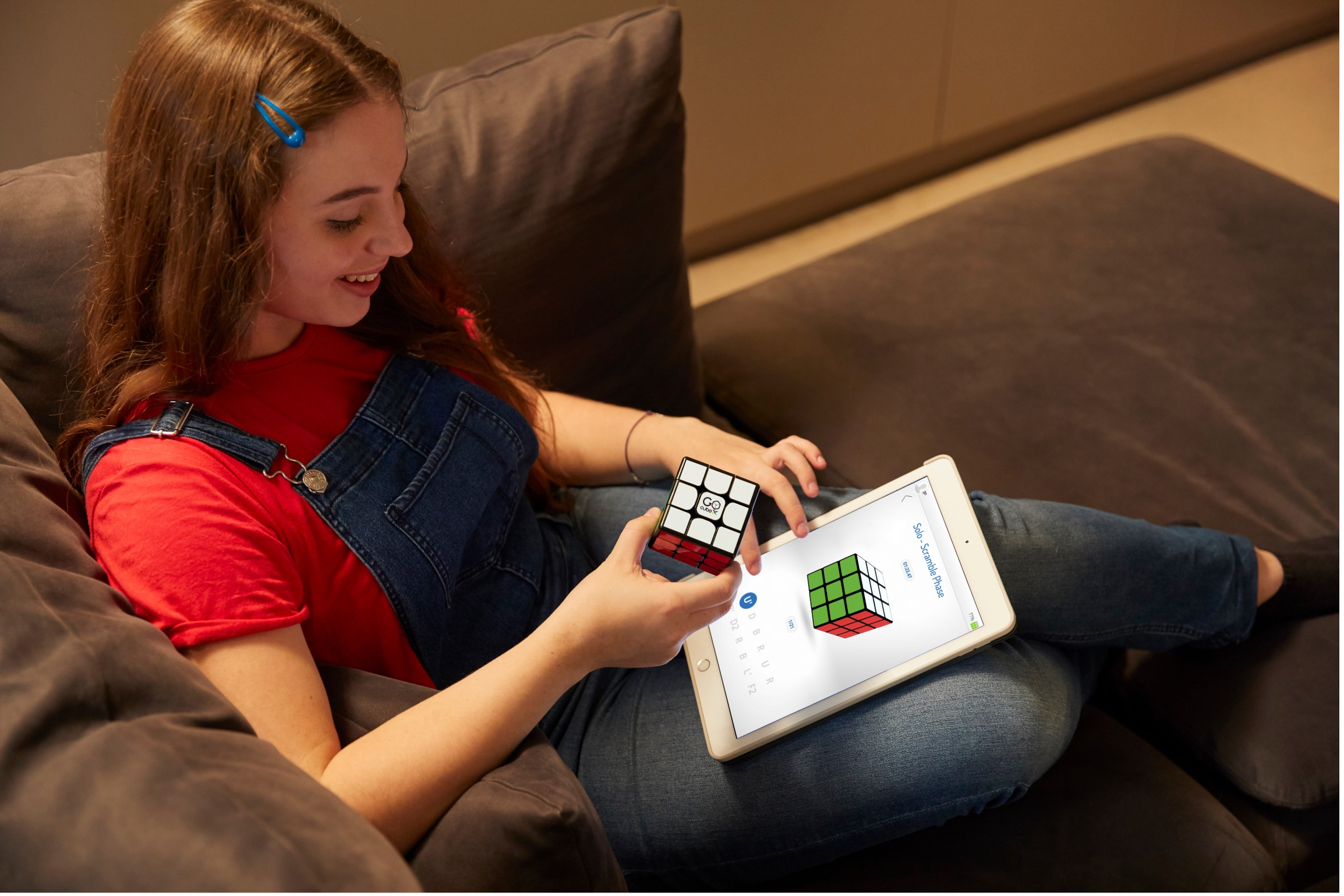 Une jeune fille assise sur un canapé tient un Rubik's cube et regarde une tablette affichant une application.