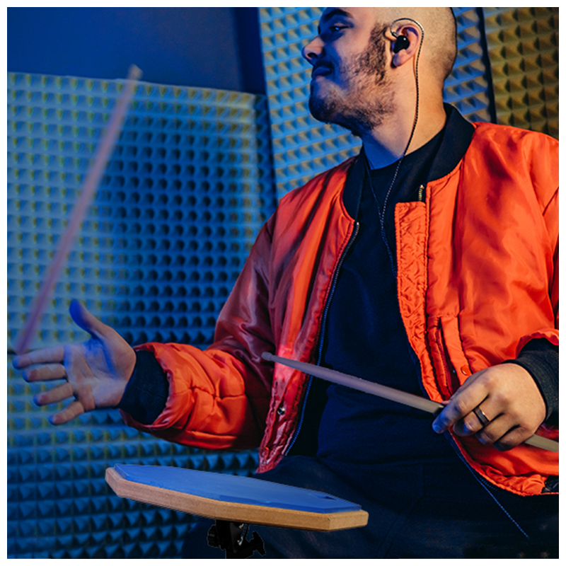 Ein Schlagzeuger in einer orangefarbenen Jacke schlägt mit Stöcken auf ein Übungspad vor Akustikschaumstoff.