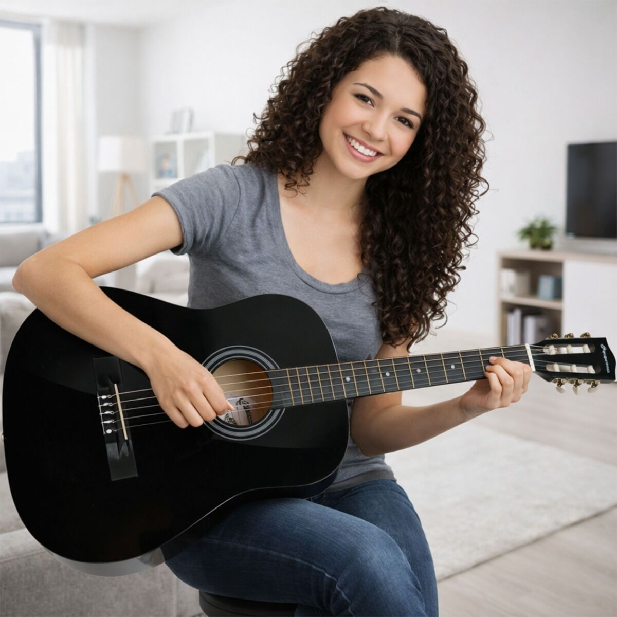 Eine lächelnde Frau mit lockigen Haaren sitzt indoors und spielt eine schwarze Akustikgitarre in einem hellen Zimmer.
