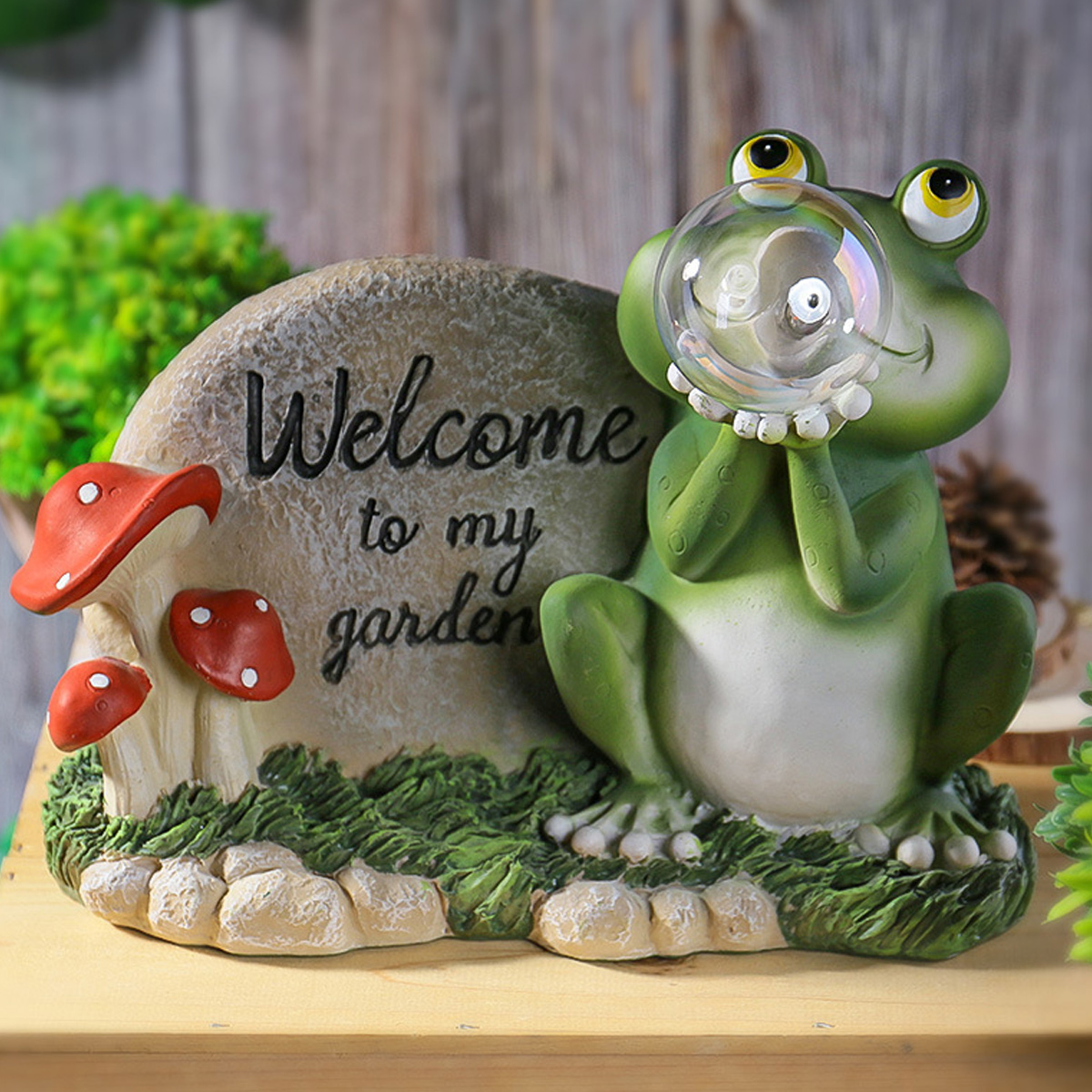 Eine grüne Frosch-Gartenstatue mit Glaskugel, neben einem Willkommen im Garten-Stein und roten Pilzen.