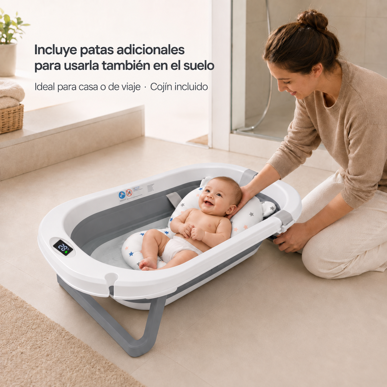 Un bebé sonriente yace en una bañera plegable gris y blanca mientras una mujer se arrodilla a su lado tocándole la cabeza.