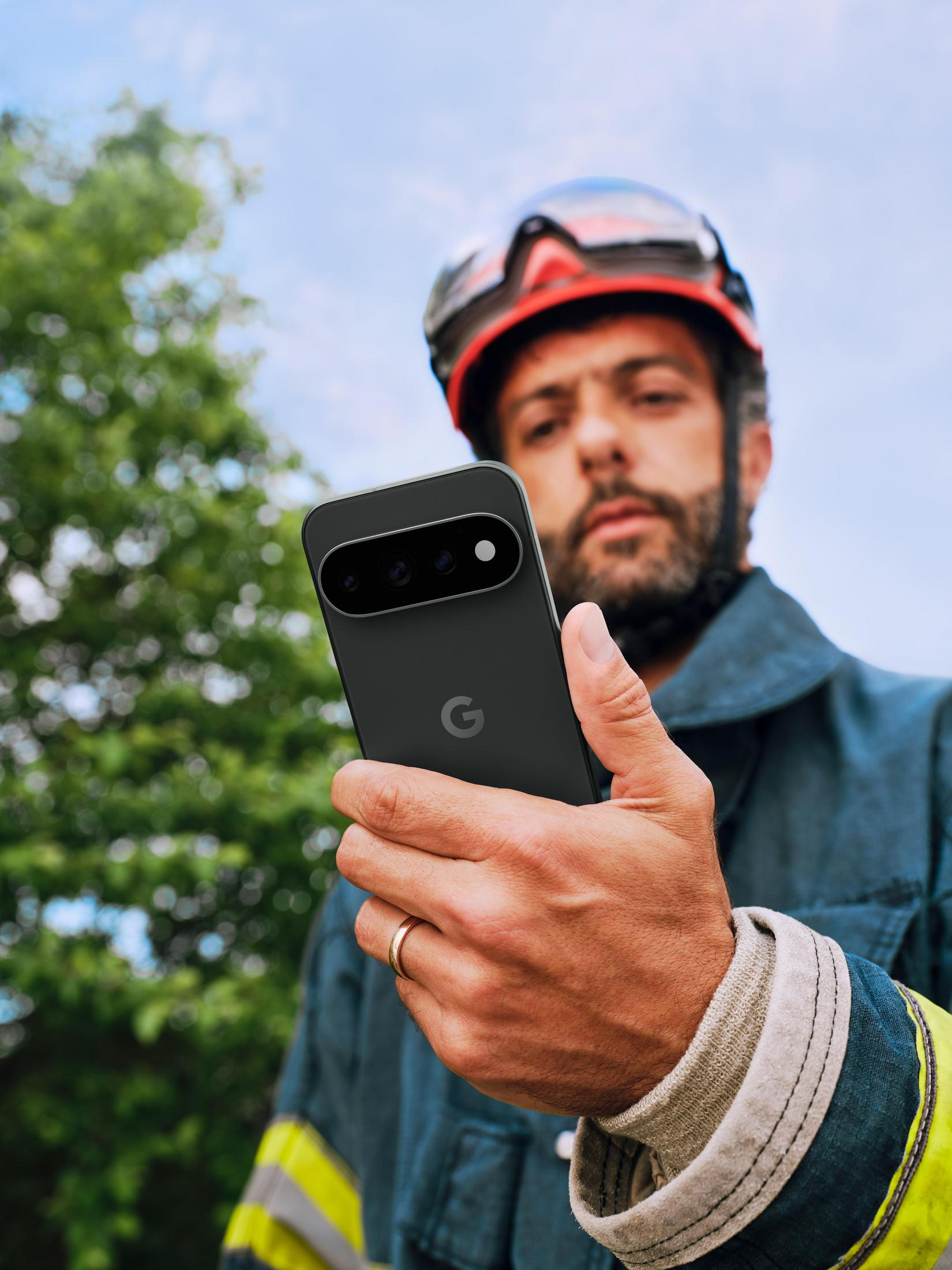 Mężczyzna w odzieży ochronnej i kasku trzyma przed sobą czarny smartfon Google Pixel.