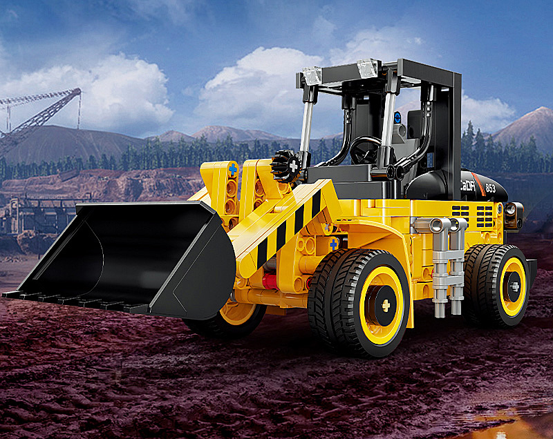 Ein gelb-schwarzer Spielzeug-Bulldozer auf schlammigem Boden vor Bergen und bewölktem Himmel.