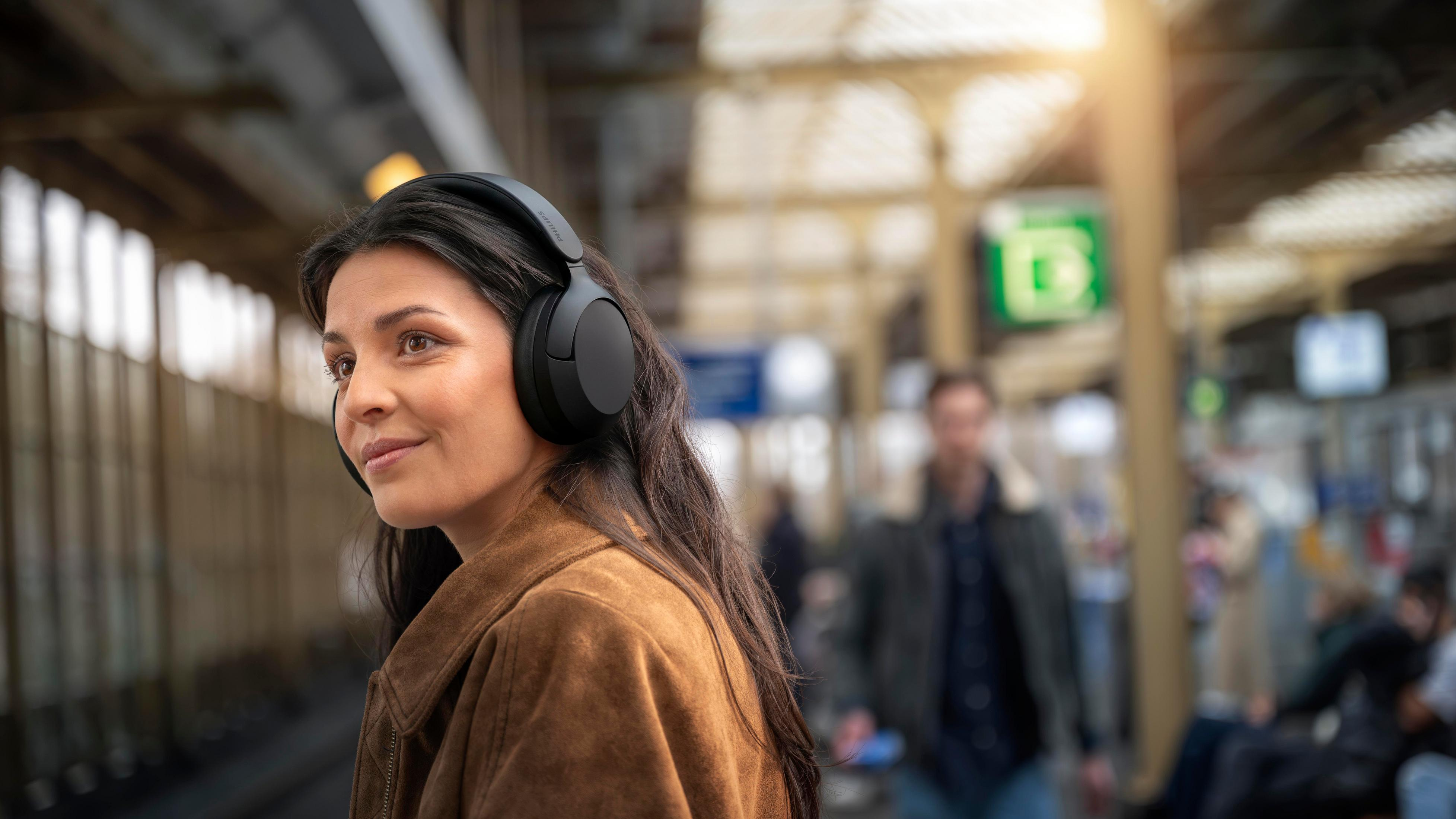 Una donna con capelli castani e cuffie si trova in una stazione ferroviaria davanti a uno sfondo sfocato.