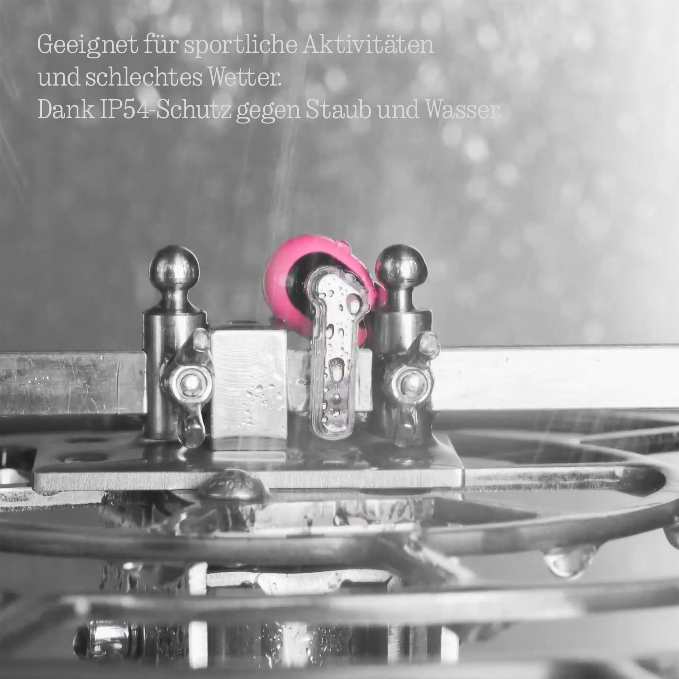 Un auricular rosa se prueba contra el agua bajo un spray con piezas mecánicas metálicas al fondo.