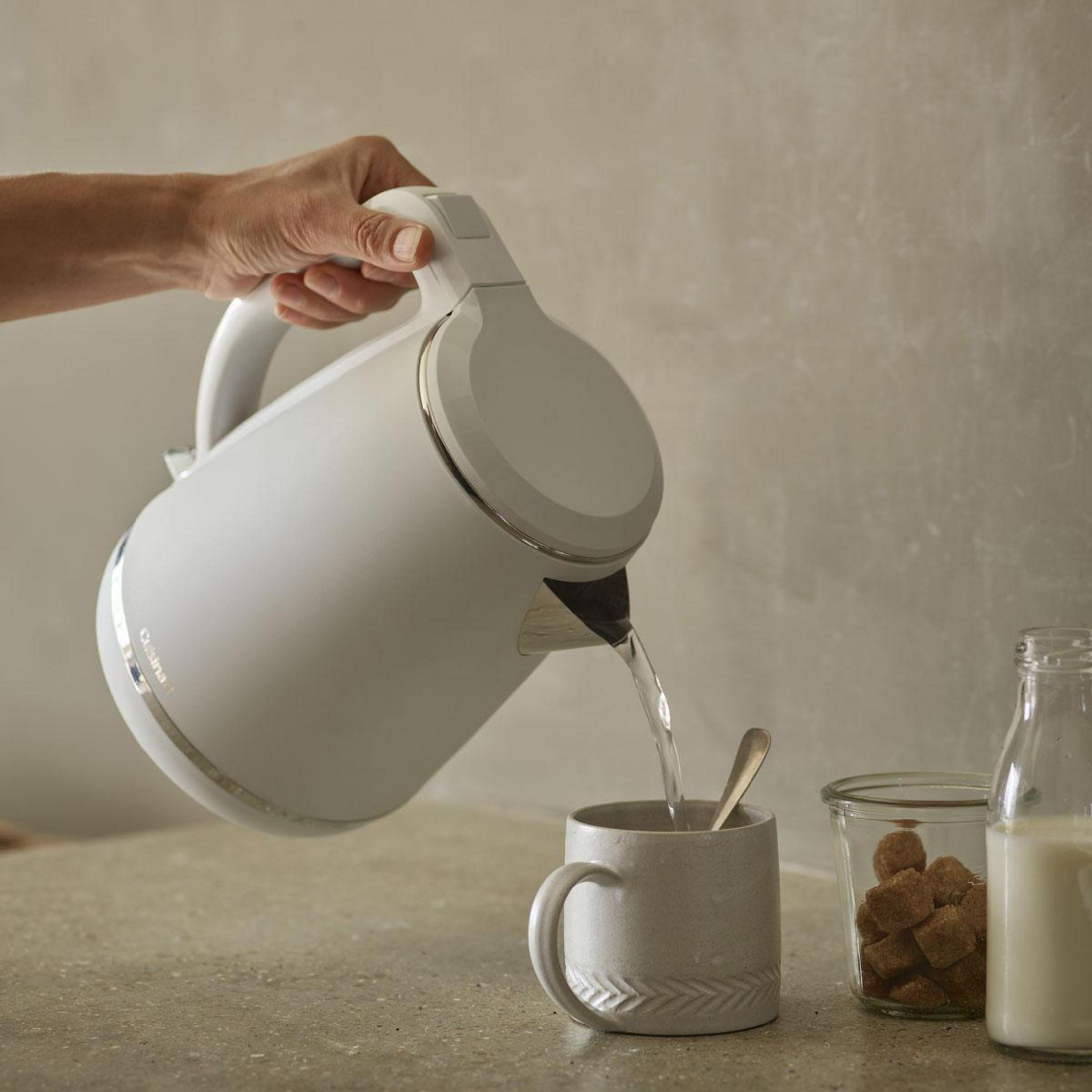 Una mano versa acqua da un bollitore elettrico bianco in una tazza di ceramica su un bancone con zucchero e latte.
