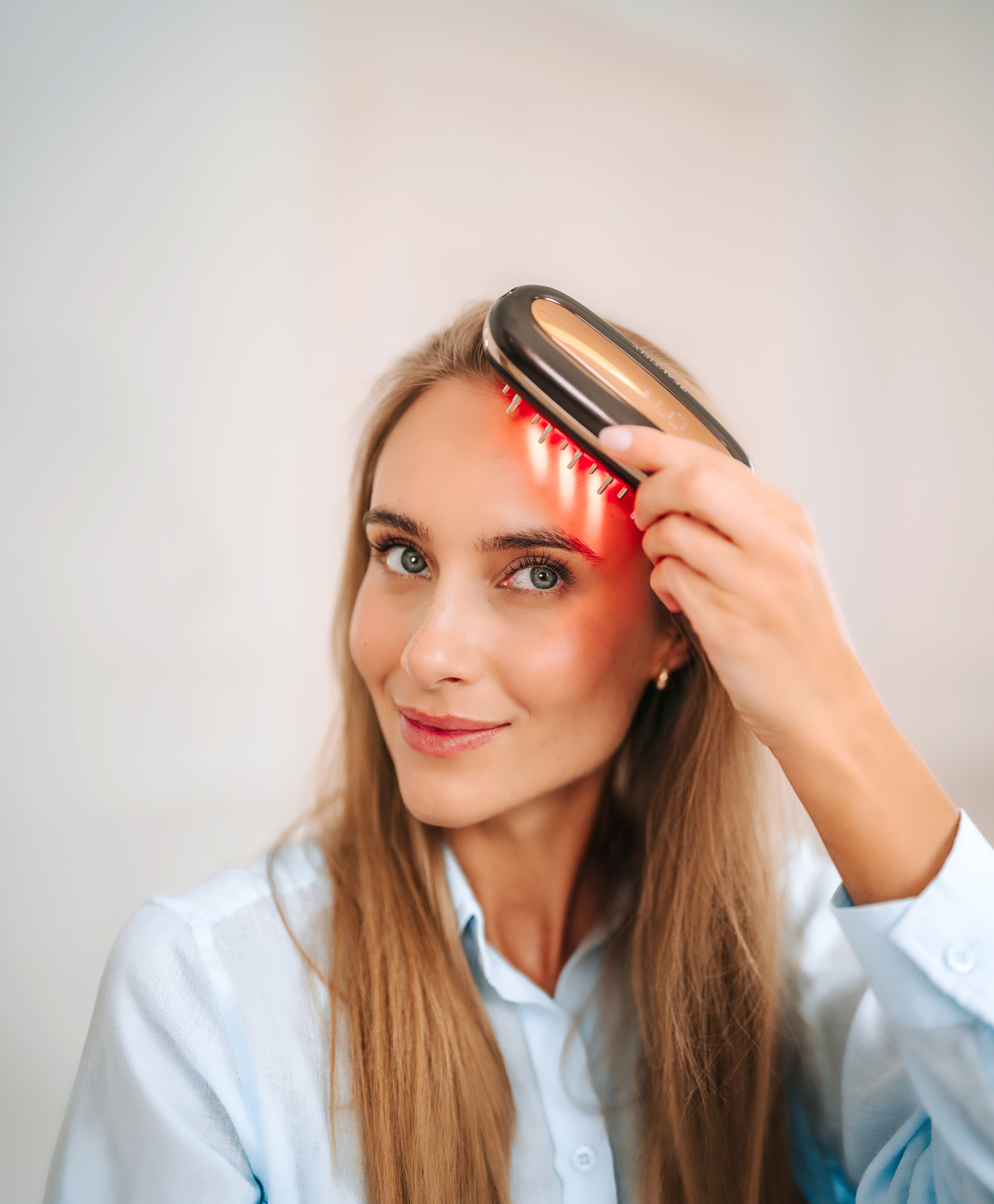 Una mujer rubia sonriente con camisa azul usa un dispositivo de terapia de luz roja en su frente.