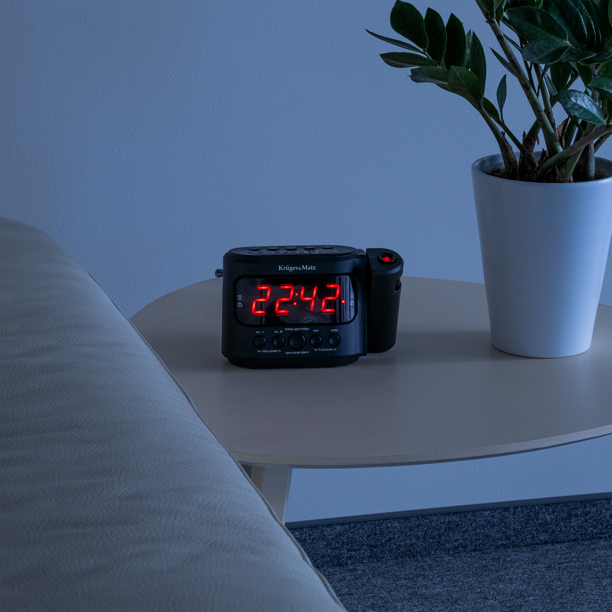 Czarny cyfrowy budzik pokazujący 22:42 na czerwono stoi na stoliku obok rośliny w doniczce.
