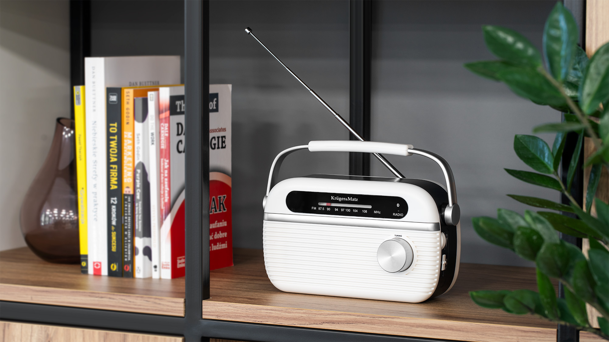 Biało-czarne radio w stylu retro z anteną i pokrętłem stoi na drewnianej półce obok książek.