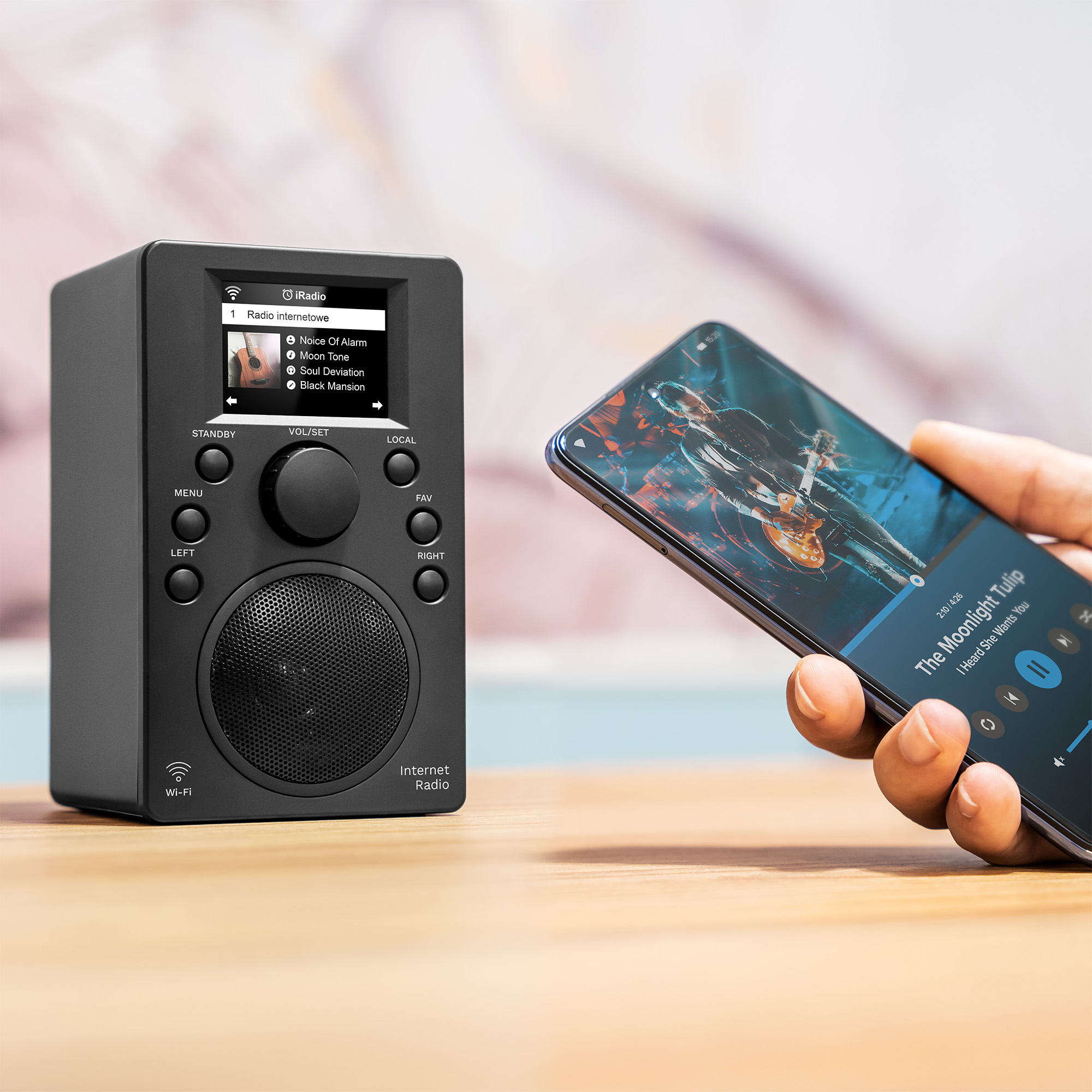 Czarne radio internetowe stoi na drewnianym stole obok dłoni trzymającej smartfon z aplikacją muzyczną.