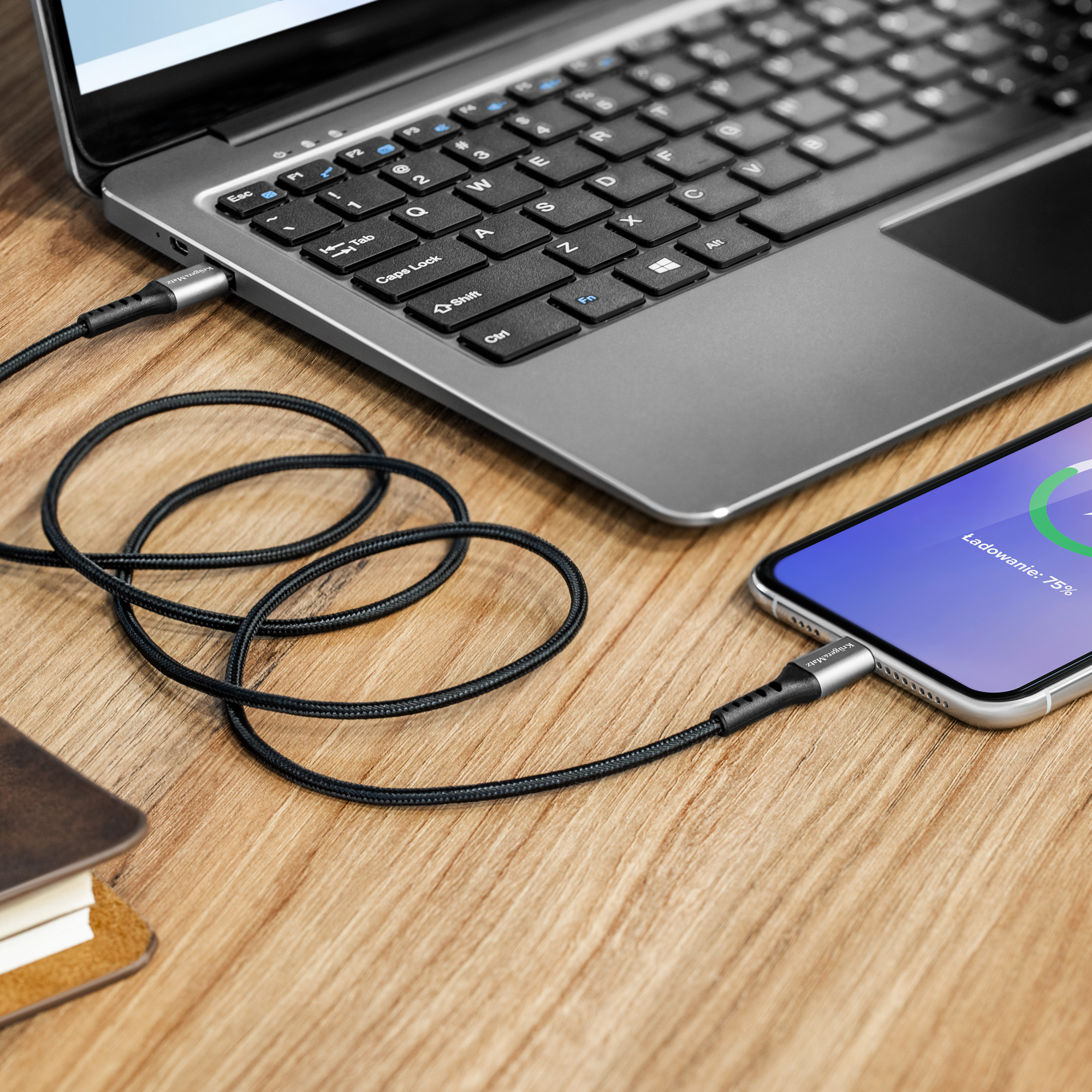 Smartfon ładuje się za pomocą czarnego kabla podłączonego do laptopa na drewnianej powierzchni.