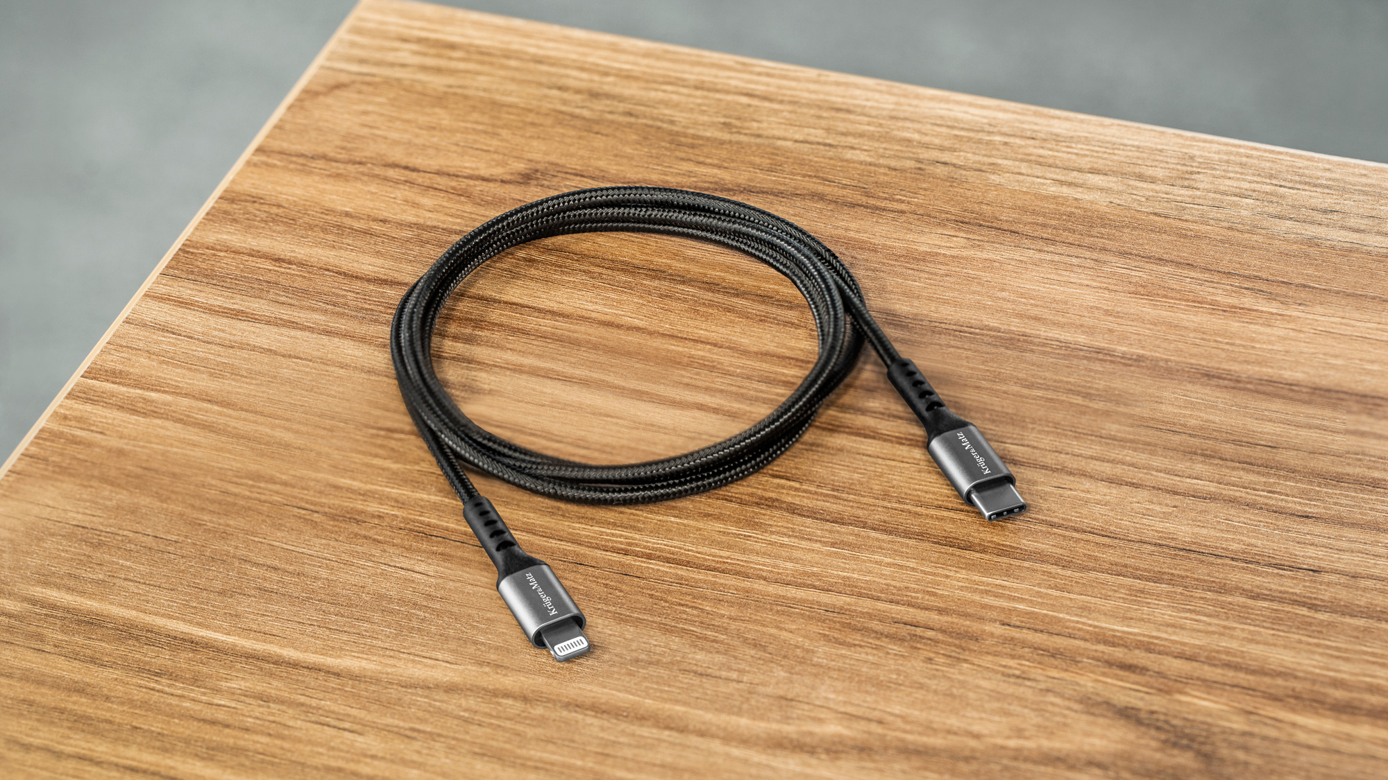 Zwinęty czarny pleciony kabel ładujący ze srebrnymi złączami USB-C i Lightning na drewnianym stole.