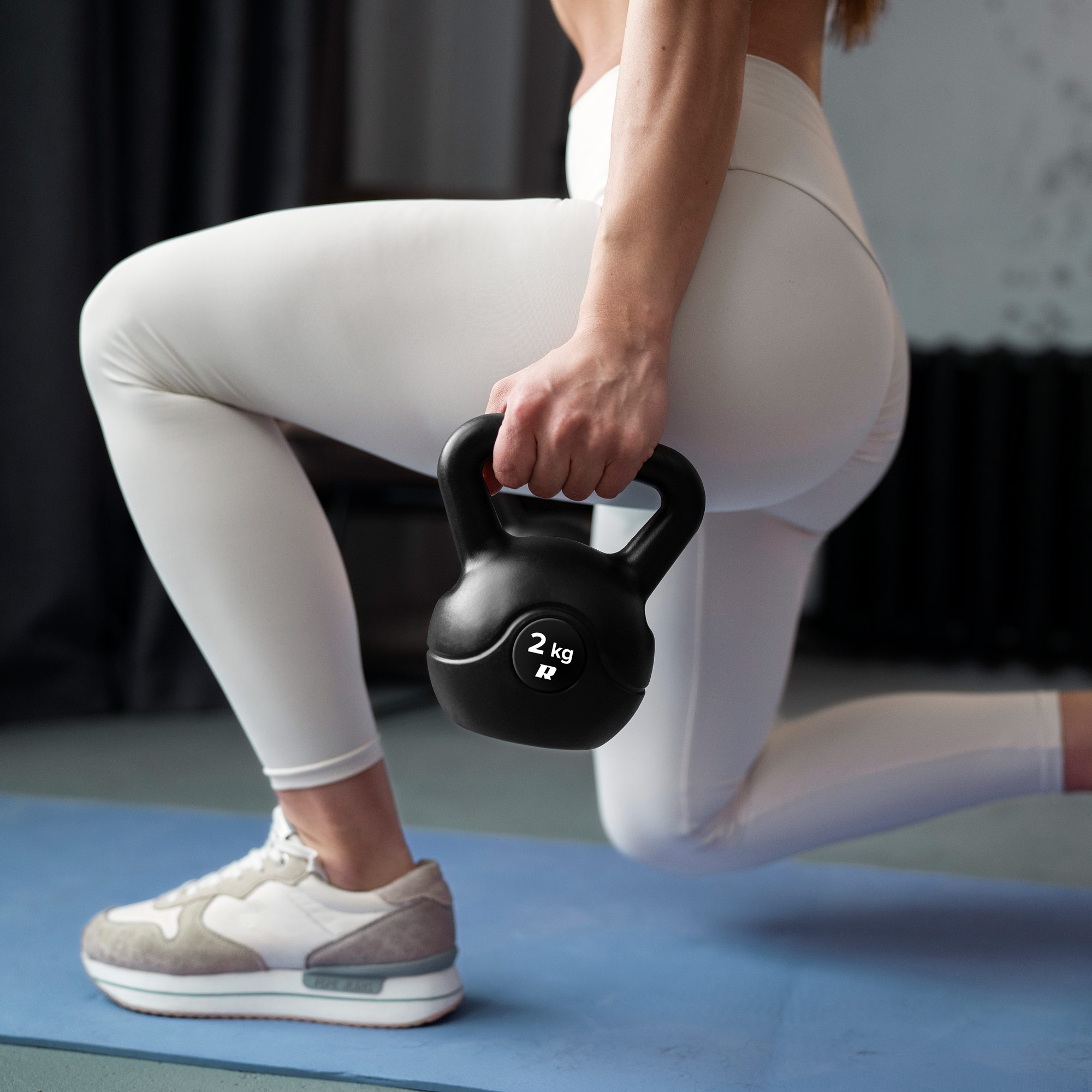 Kobieta w białych legginsach robi wykroki na niebieskiej macie, trzymając czarny kettlebell 2 kg.
