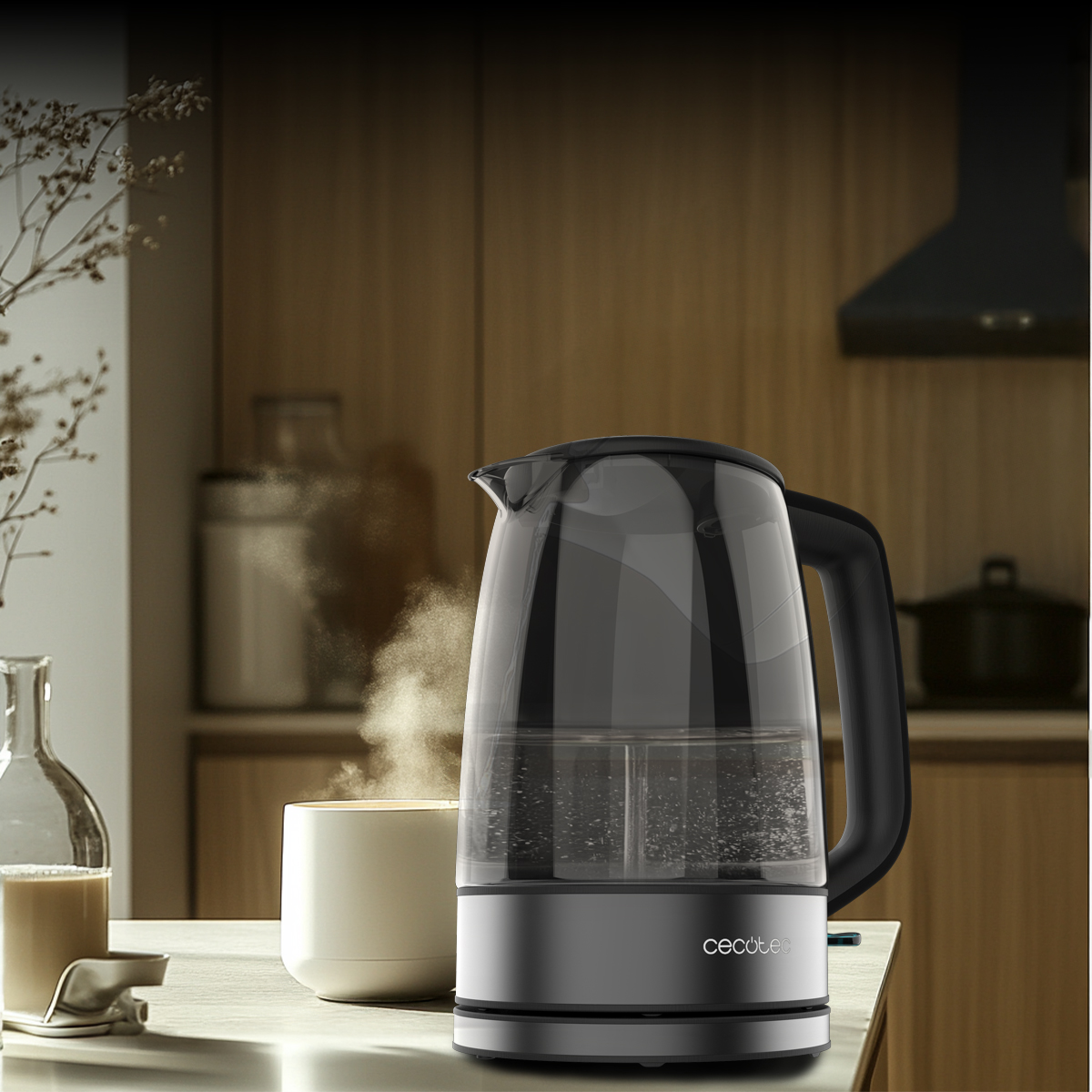 Un hervidor de vidrio con asa negra se encuentra en la encimera de una cocina junto a una taza humeante.
