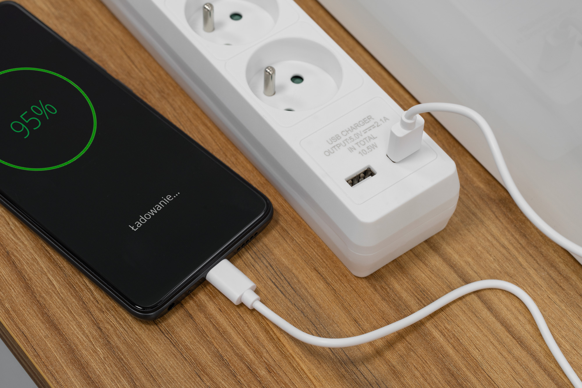 Smartfon z baterią 95% ładuje się przez białą ładowarkę USB podłączoną do listwy zasilającej.
