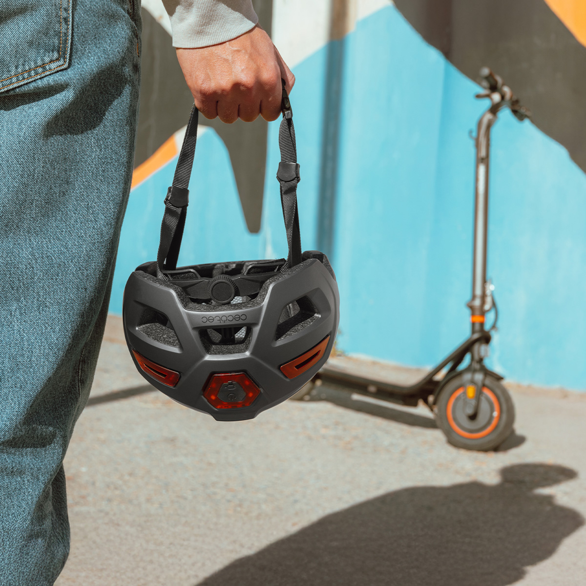 Una mano sostiene un casco negro, con un patinete eléctrico aparcado contra una pared colorida.