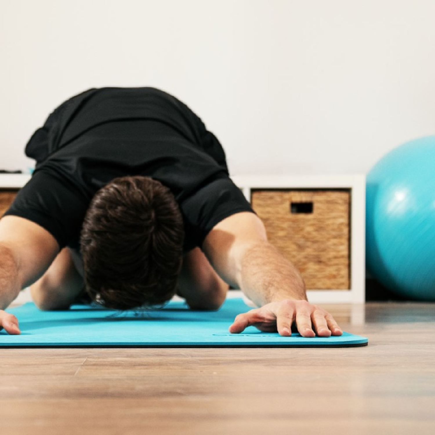 Un uomo in maglietta nera fa stretching su un tappetino da yoga blu, con palla da ginnastica blu e pavimento in legno.