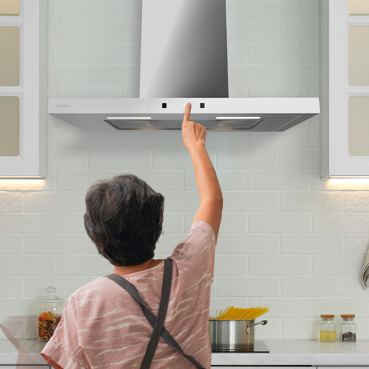 Una mujer con delantal opera el panel táctil de una campana extractora moderna en una cocina blanca.