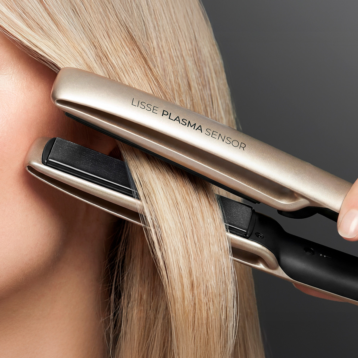 Piastra per capelli dorata modella capelli biondi su sfondo grigio.