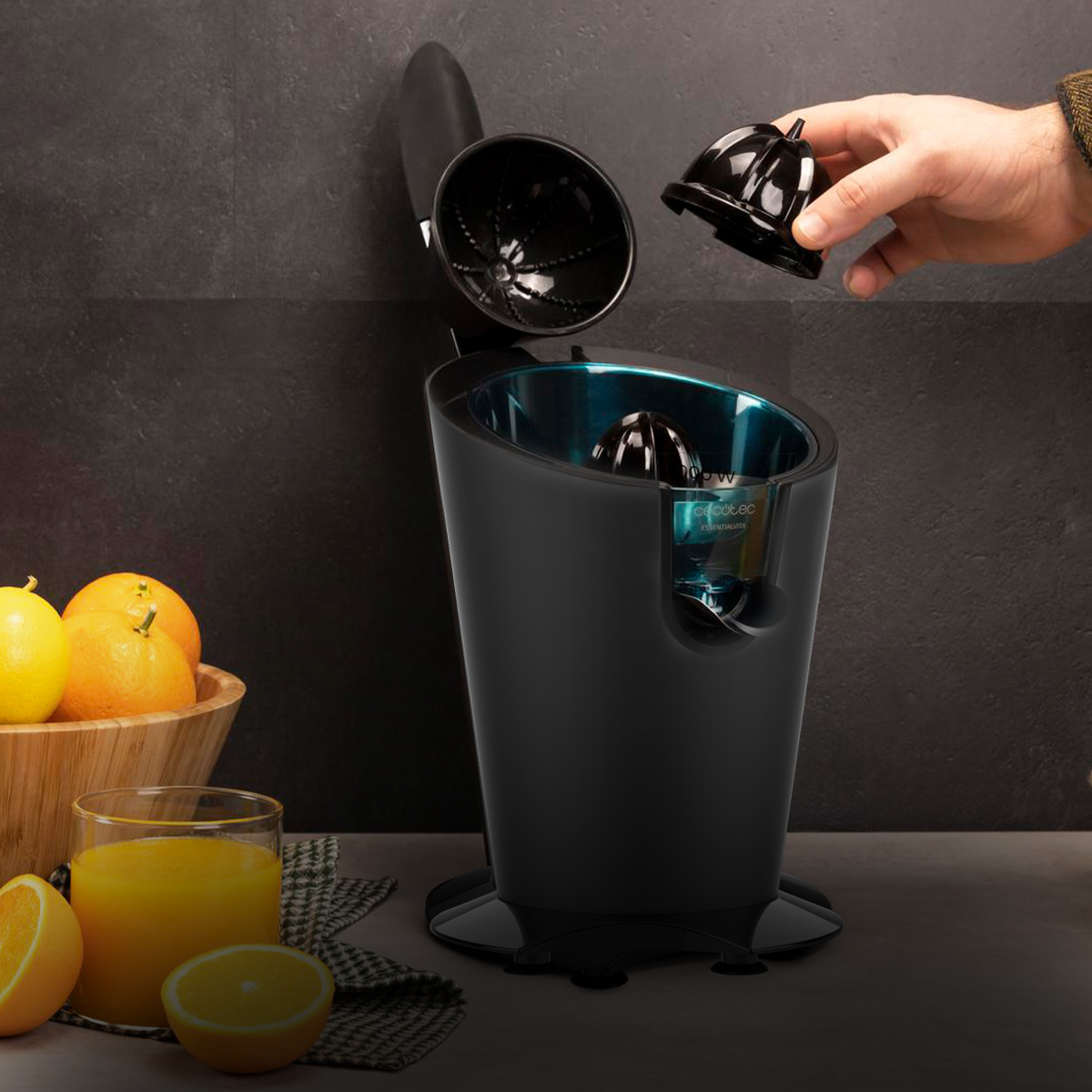 Mano inserisce un cono in uno spremiagrumi elettrico nero accanto a una ciotola di arance e un bicchiere di succo.