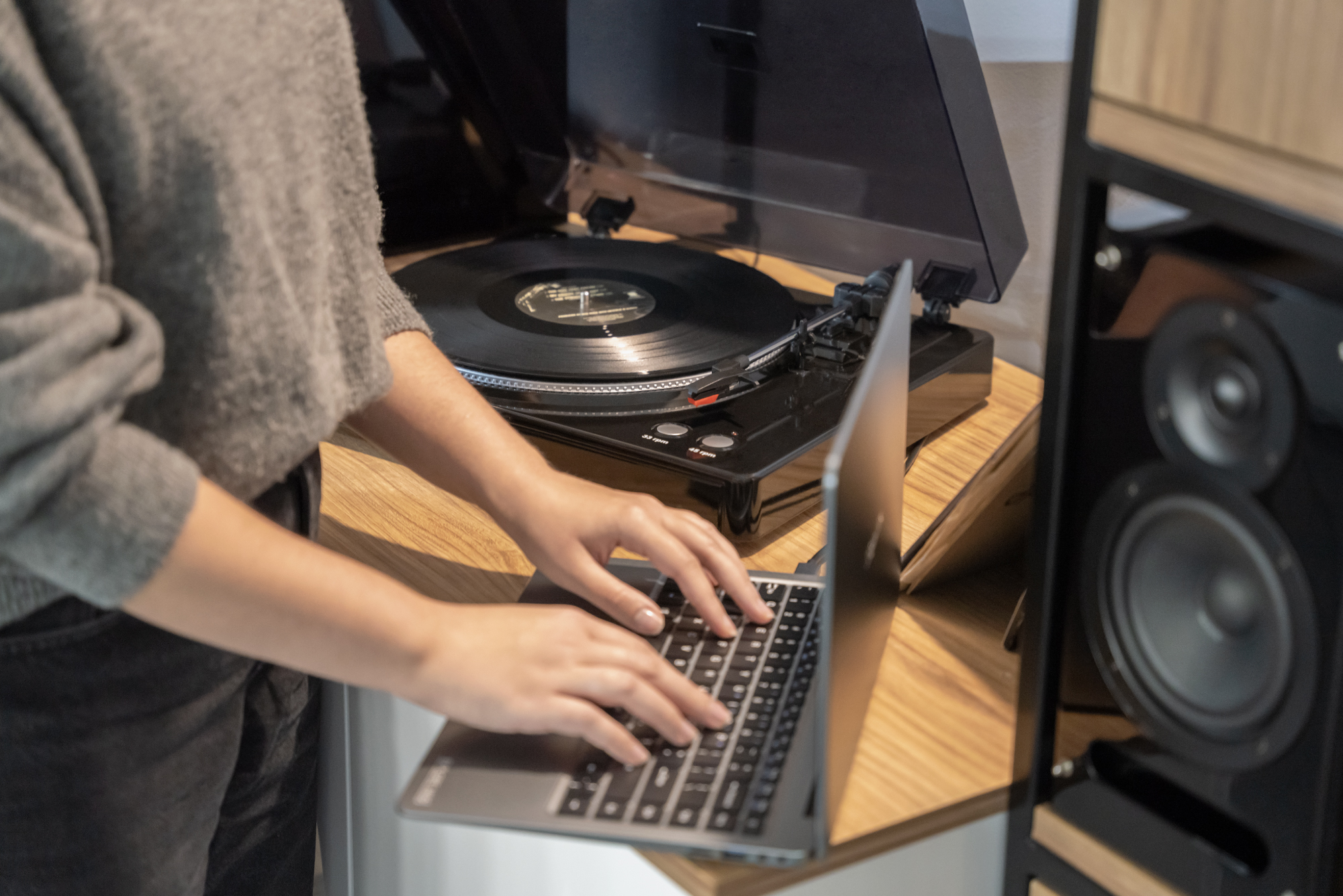 Kobieta w szarym swetrze pisze na laptopie, w tle znajduje się gramofon na drewnianej powierzchni.