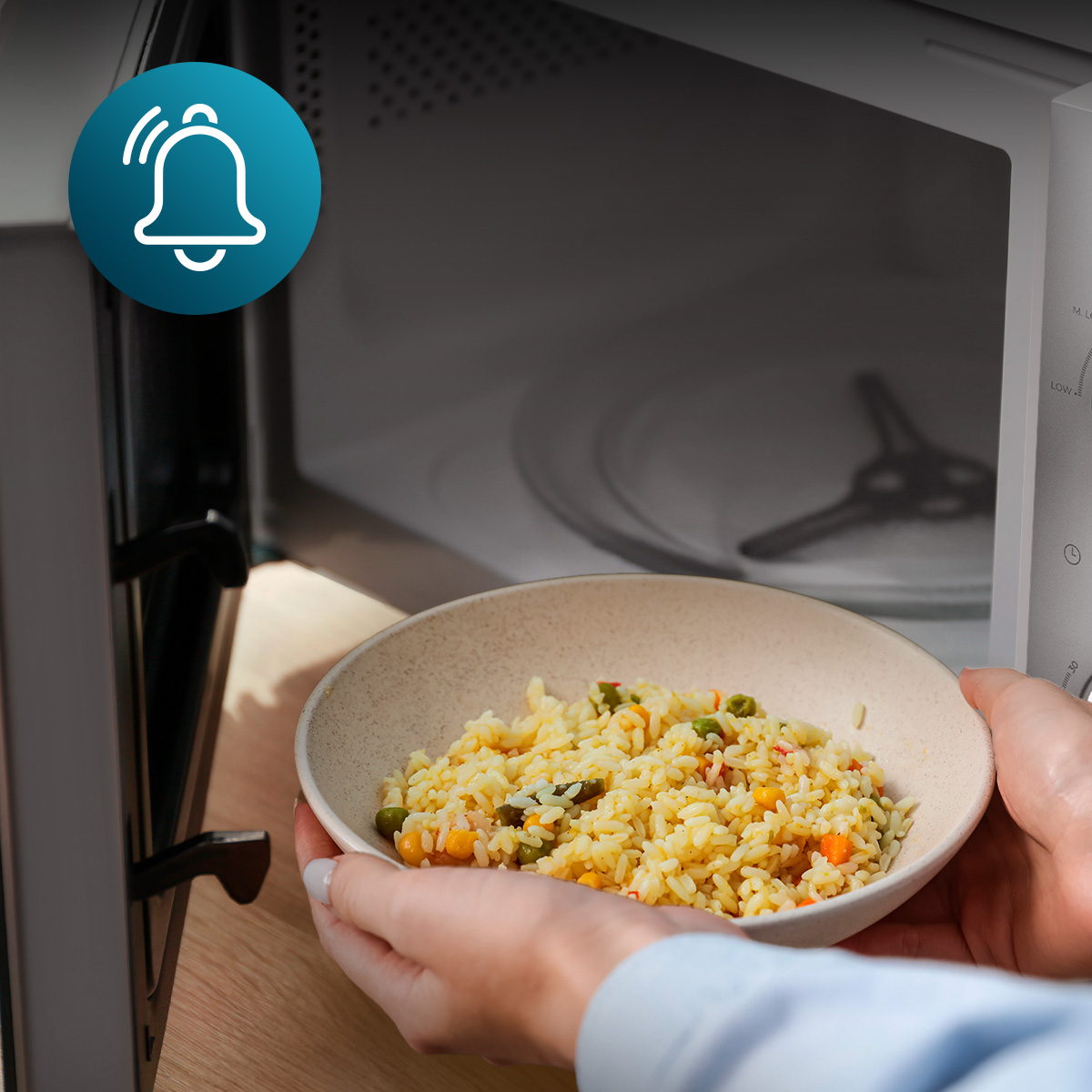 Una persona sostiene un tazón de arroz con verduras frente a un microondas abierto, con un icono de campana.