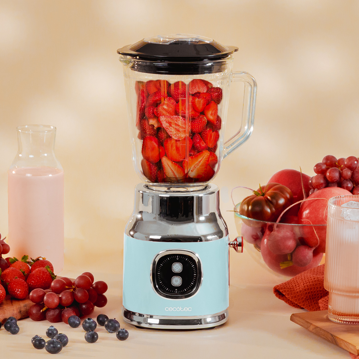 Una licuadora azul claro y cromada llena de fresas picadas sobre una mesa con fruta, leche y un vaso de batido.