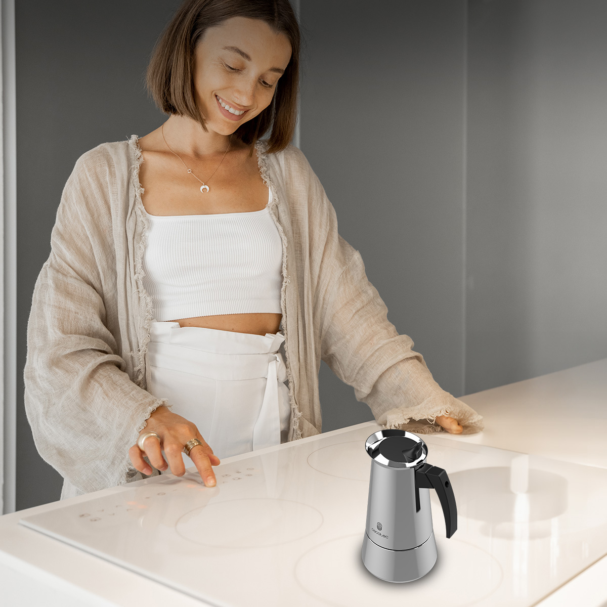 Una donna con abiti chiari aziona un piano cottura a induzione bianco con una caffettiera argentata.